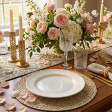 Lade das Bild in den Galerie-Viewer, A styled dining table setting with a large floral centerpiece in pink and white roses, gold candlesticks, clear textured glassware, woven placemats, and white plates on a wooden table.
